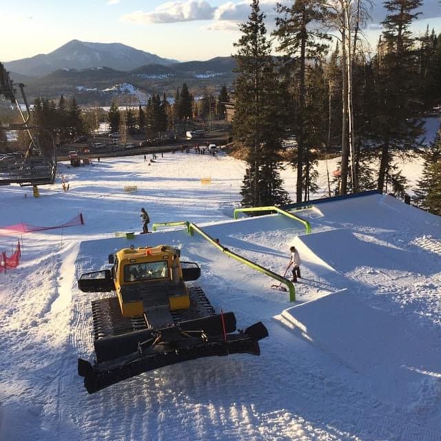 A yellow snowcat parked above a freshly built rail garden with green rails, a crew member raking the landing and a mountain in the background