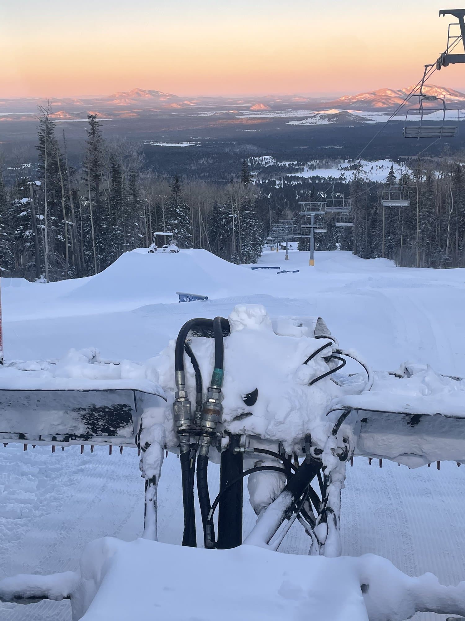 Looking over the snow-caked hydraulic lines on the back of a snowcat toward a freshly built park jump, with sunrise lighting up the desert peaks in the distance