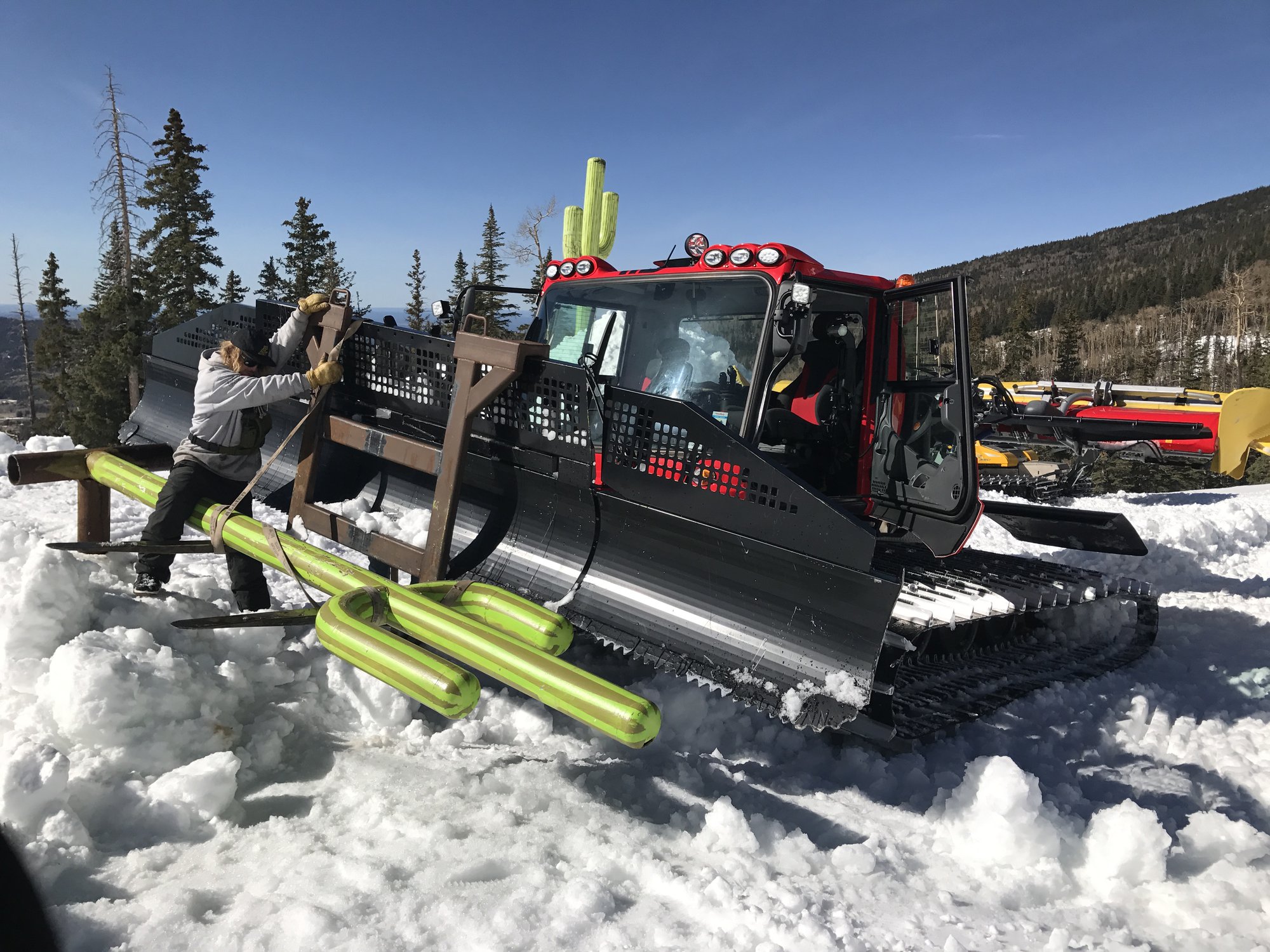 A park crew member ratchet-strapping a green cactus-shaped rail feature to the blade of a red snowcat on a bluebird day