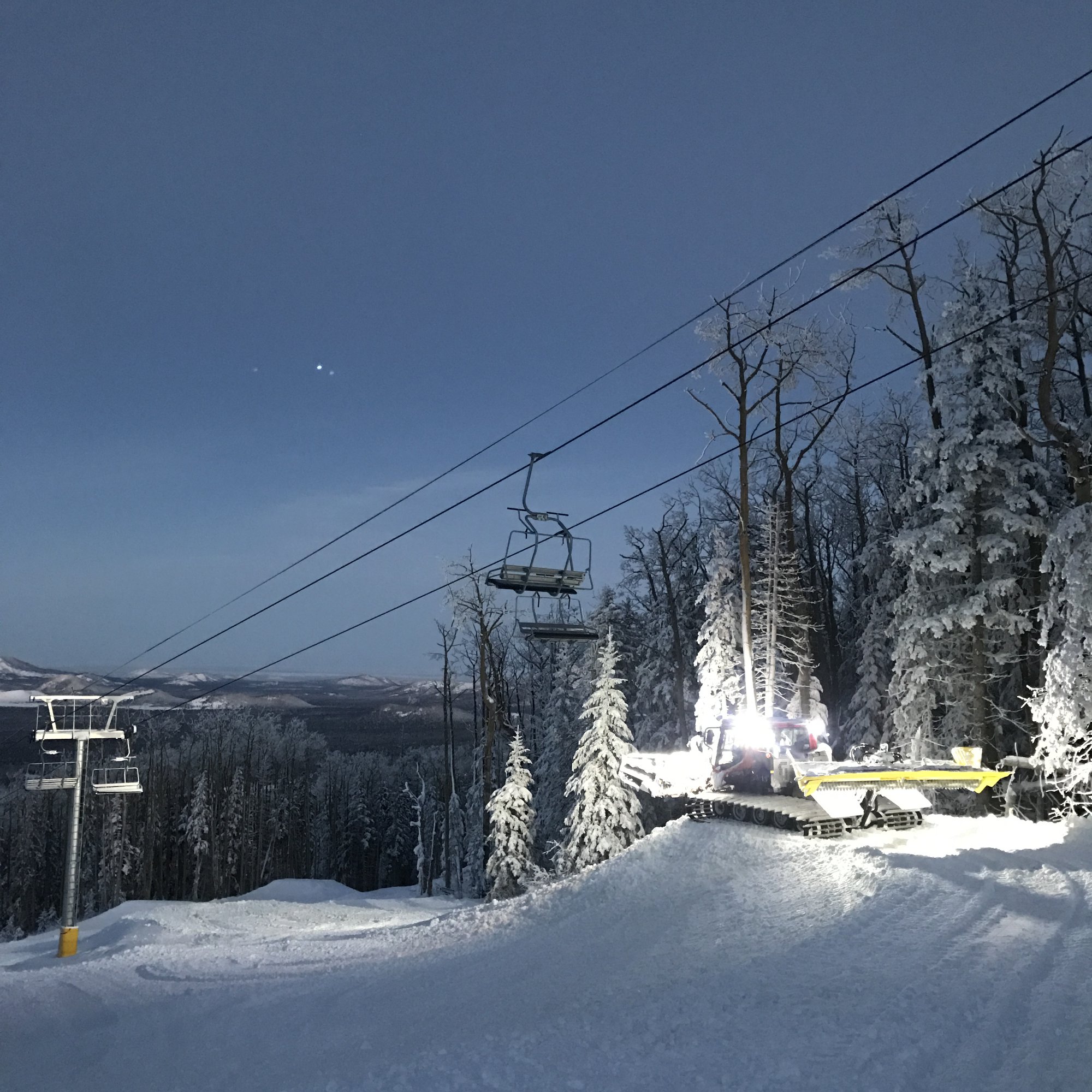 A snowcat lit up at dusk beneath a chairlift, pushing snow for a park build with frosted trees behind it