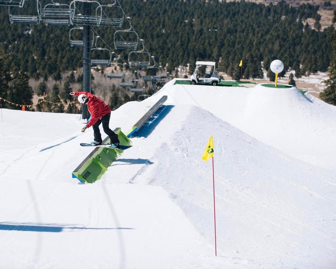 A snowboarder in a red jacket boardslides a green box below a golf-themed park feature with a golf cart and giant golf ball