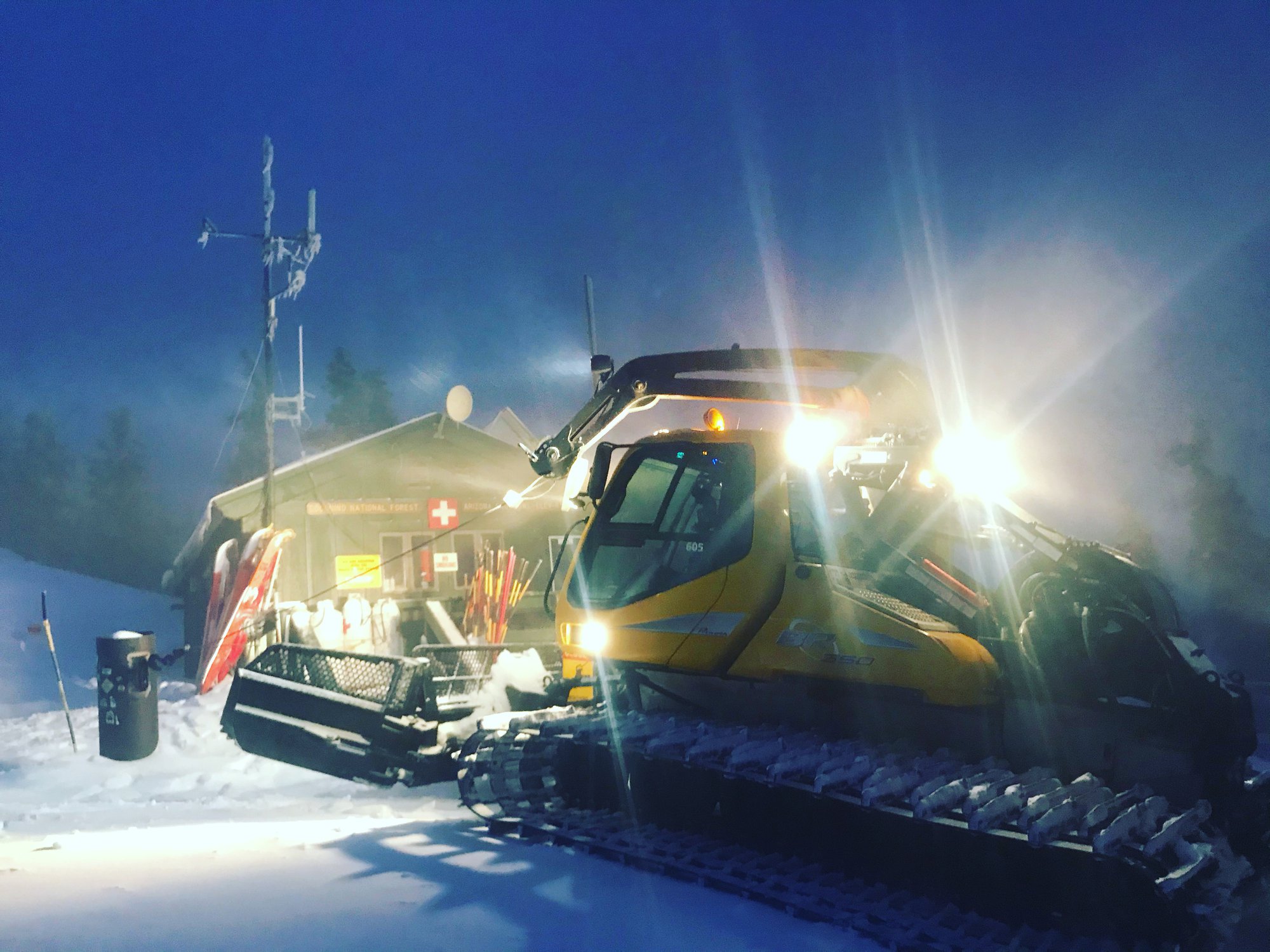 A yellow snowcat with its work lights blazing in blowing snow in front of a ski patrol shack at night