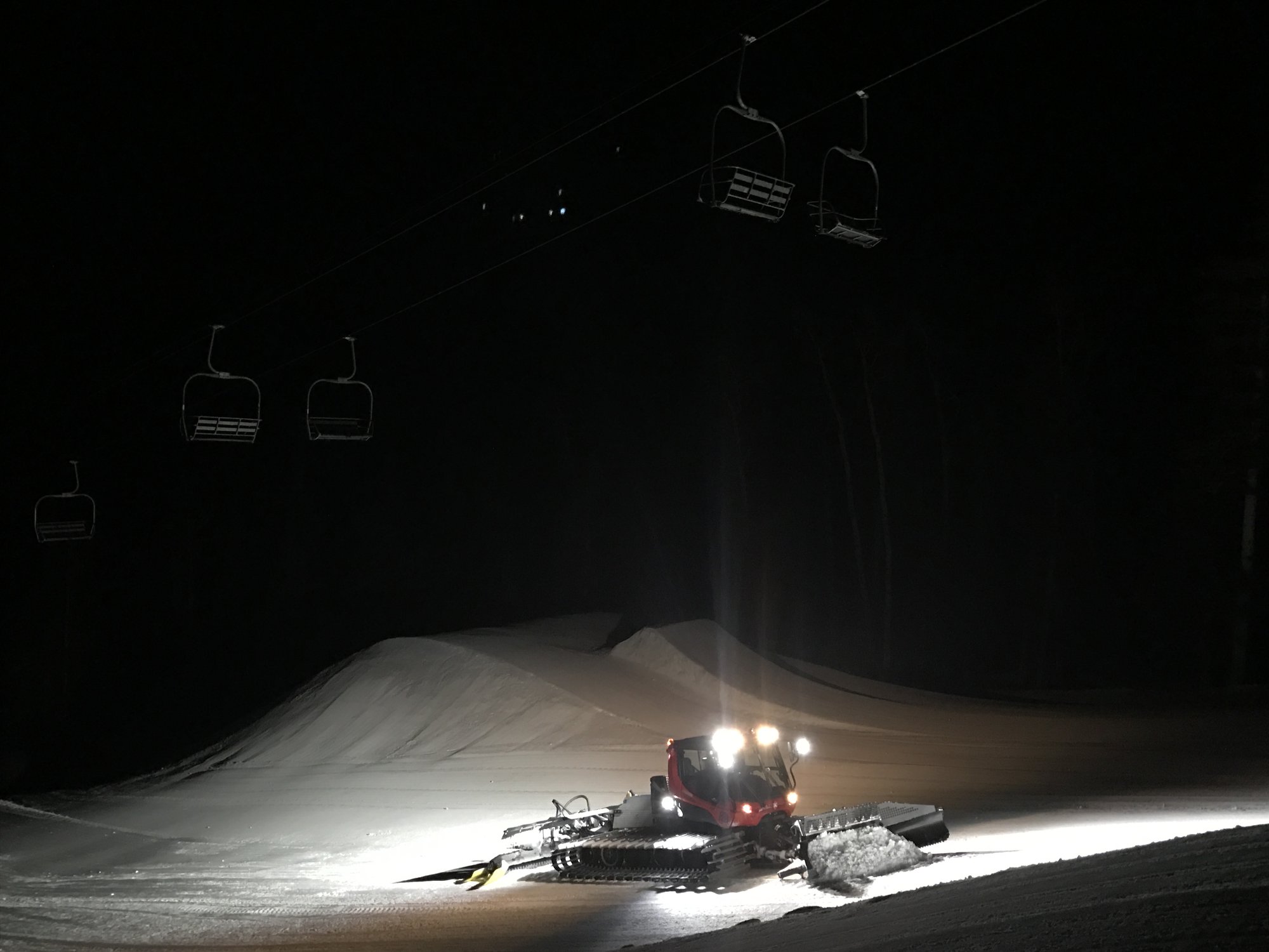 A red snowcat with its lights on shaping a line of jumps at night beneath an empty chairlift
