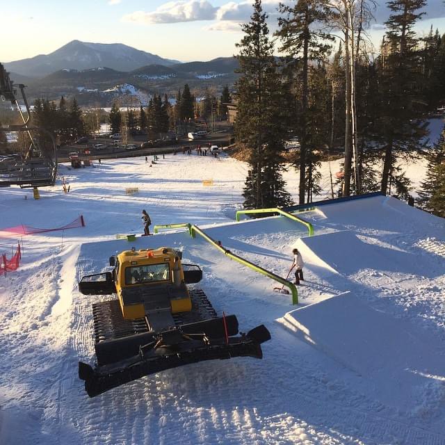 A yellow snowcat parked above a freshly built rail garden with green rails, a crew member raking the landing and a mountain in the background