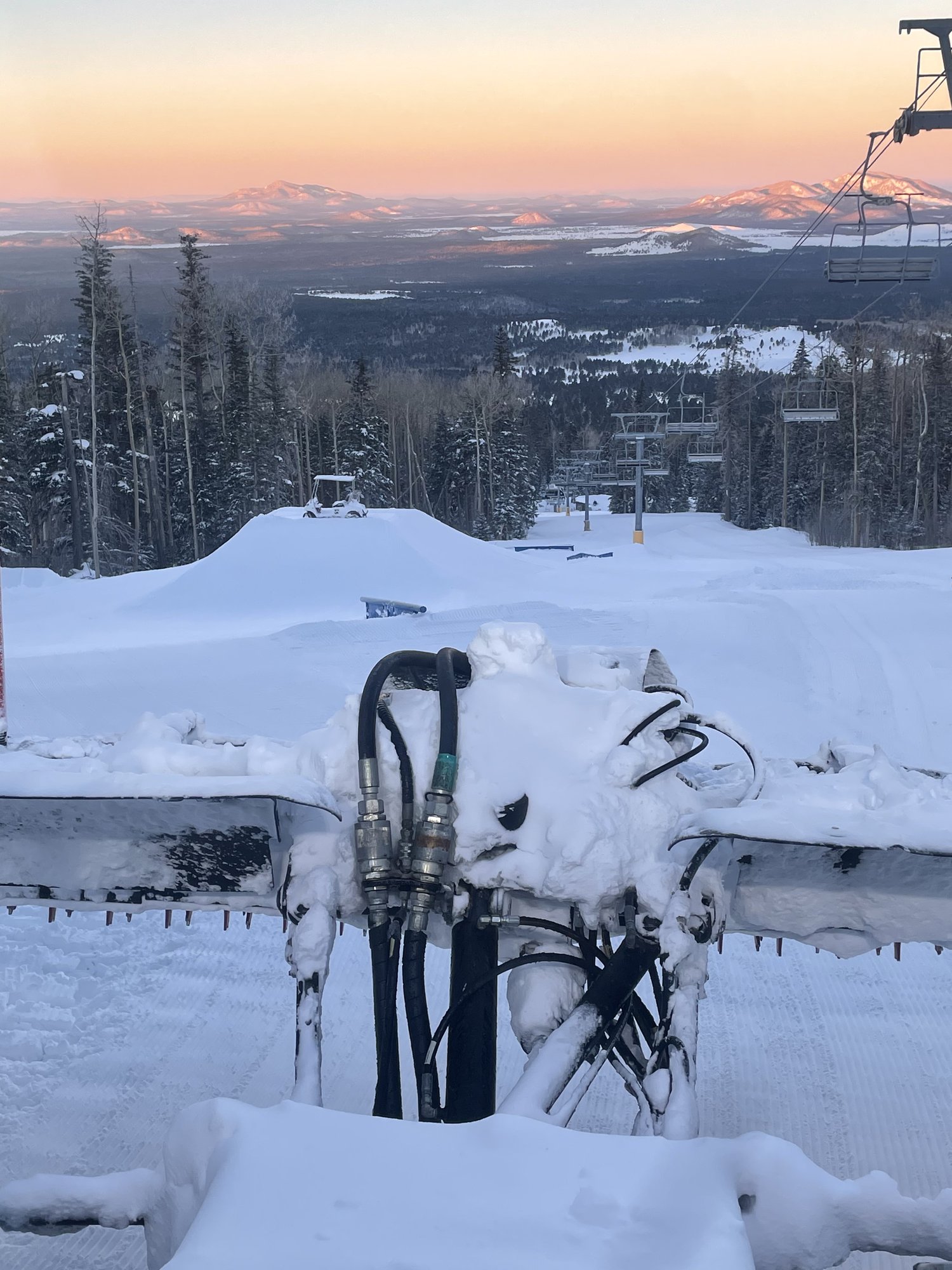 Looking over the snow-caked hydraulic lines on the back of a snowcat toward a freshly built park jump, with sunrise lighting up the desert peaks in the distance